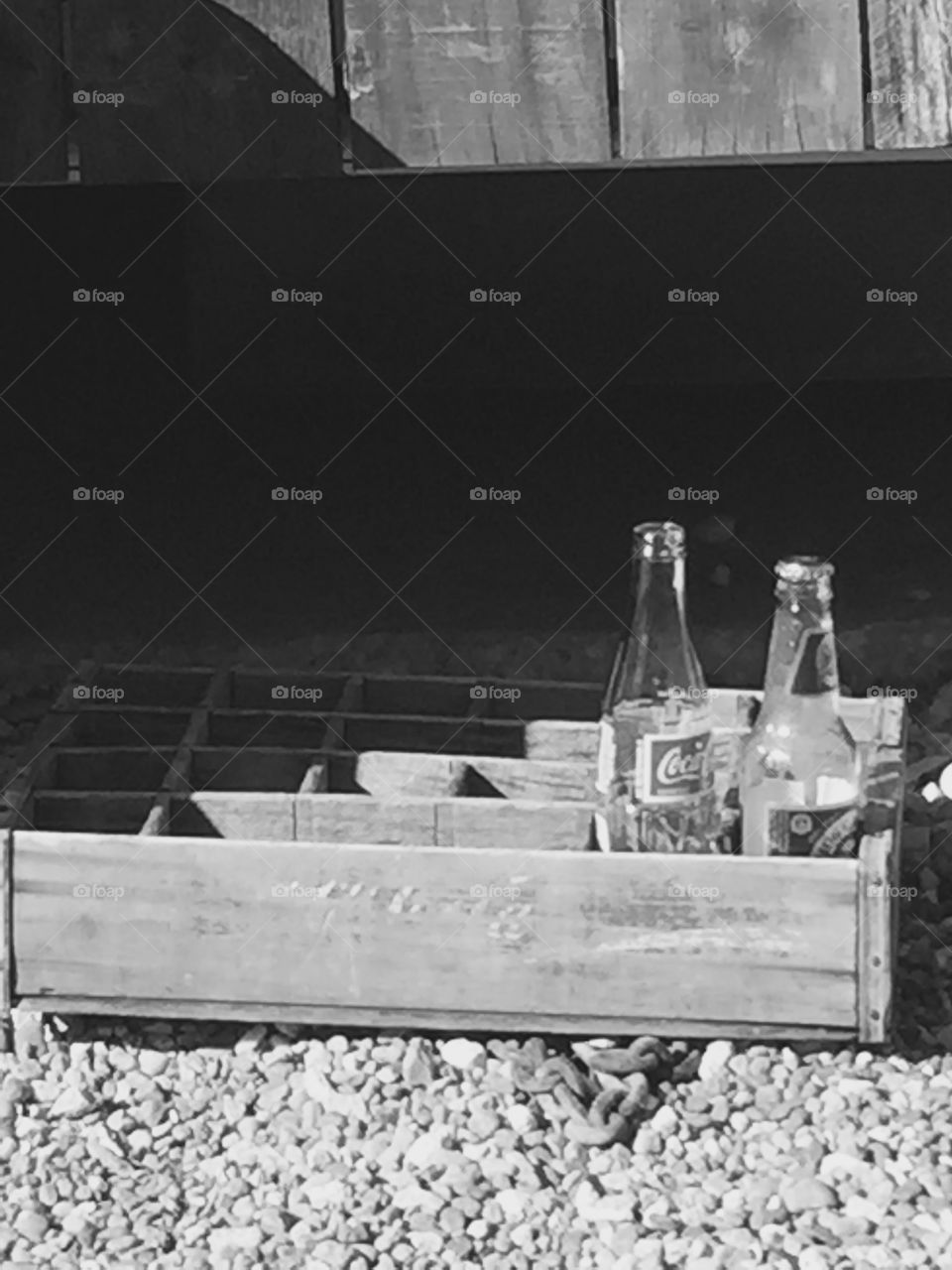 Black and white photo of a wooden grate on a gravel ground and two empty glass soda bottles inside
