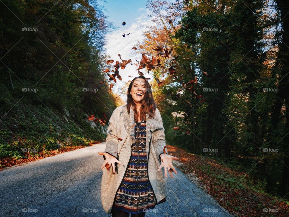 Happy woman standing on road, throwing autumn leaves in air