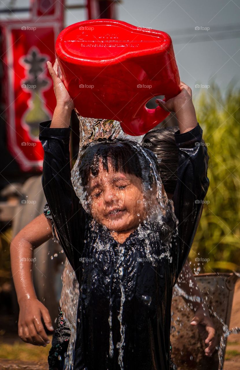 A young child joyfully pours water over their head from a red container, capturing a refreshing moment of play under the sun. The expression on the child's face reflects pure bliss, with water streaming down, adding a sense of movement and energy t