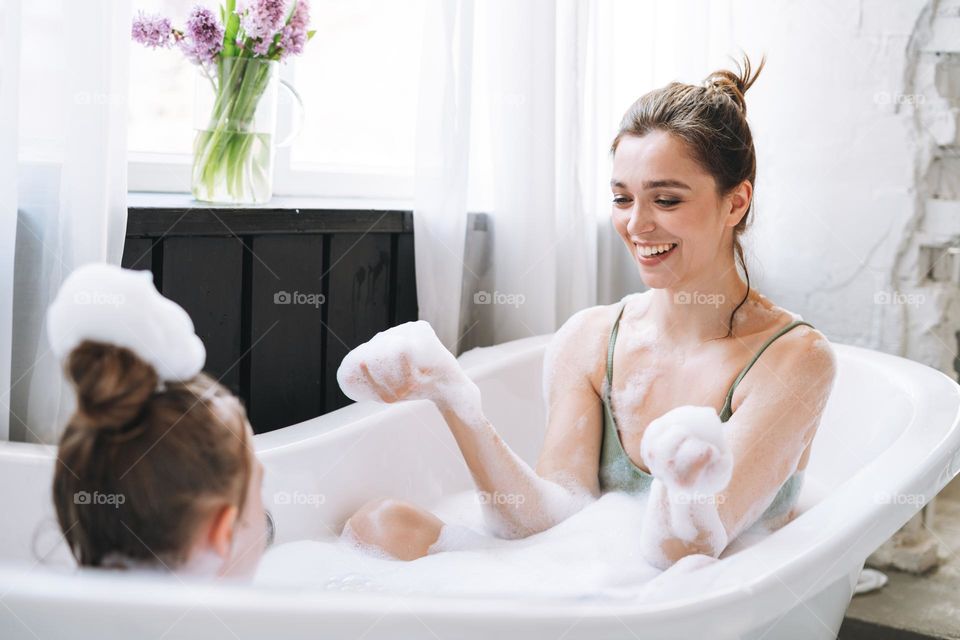 Young mother woman and little tween girl daughter having fun in bath with foam at home