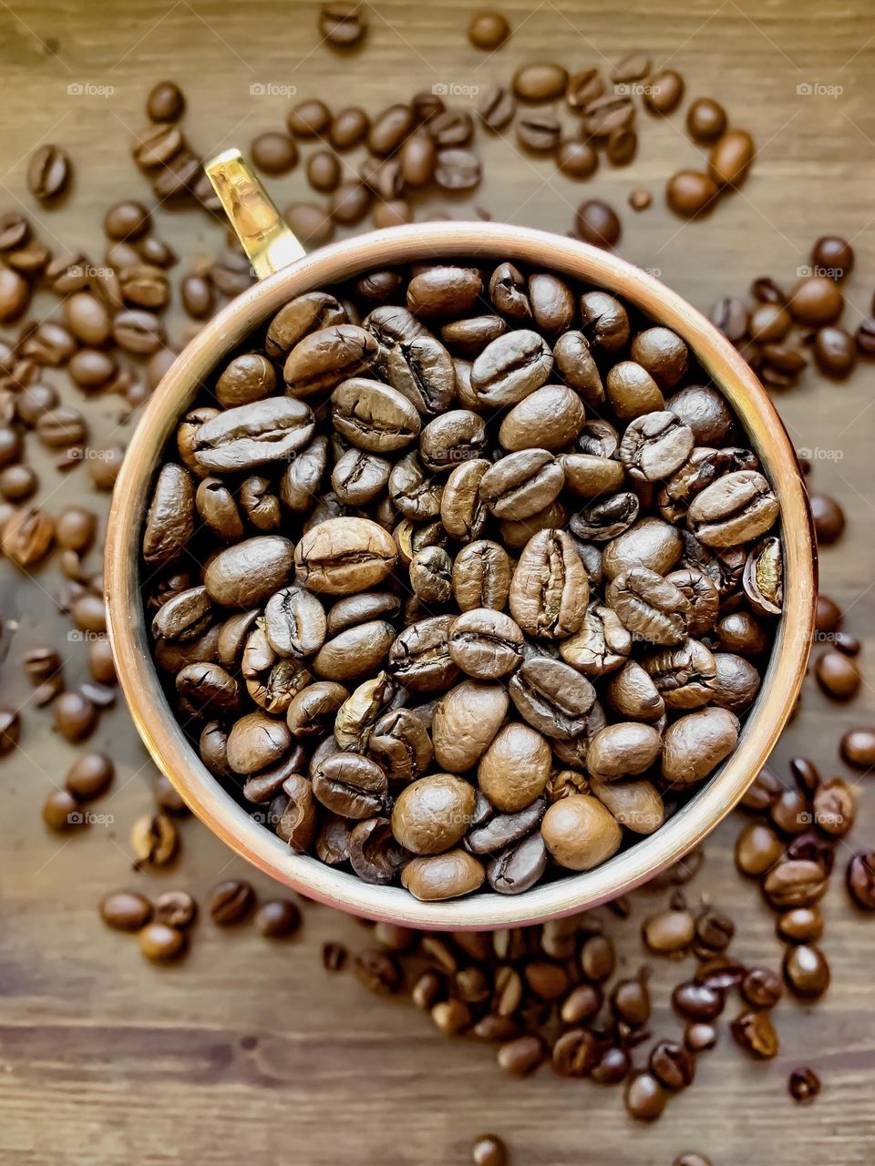 A copper mug full of coffee beans with more beans around it on a wooden background 