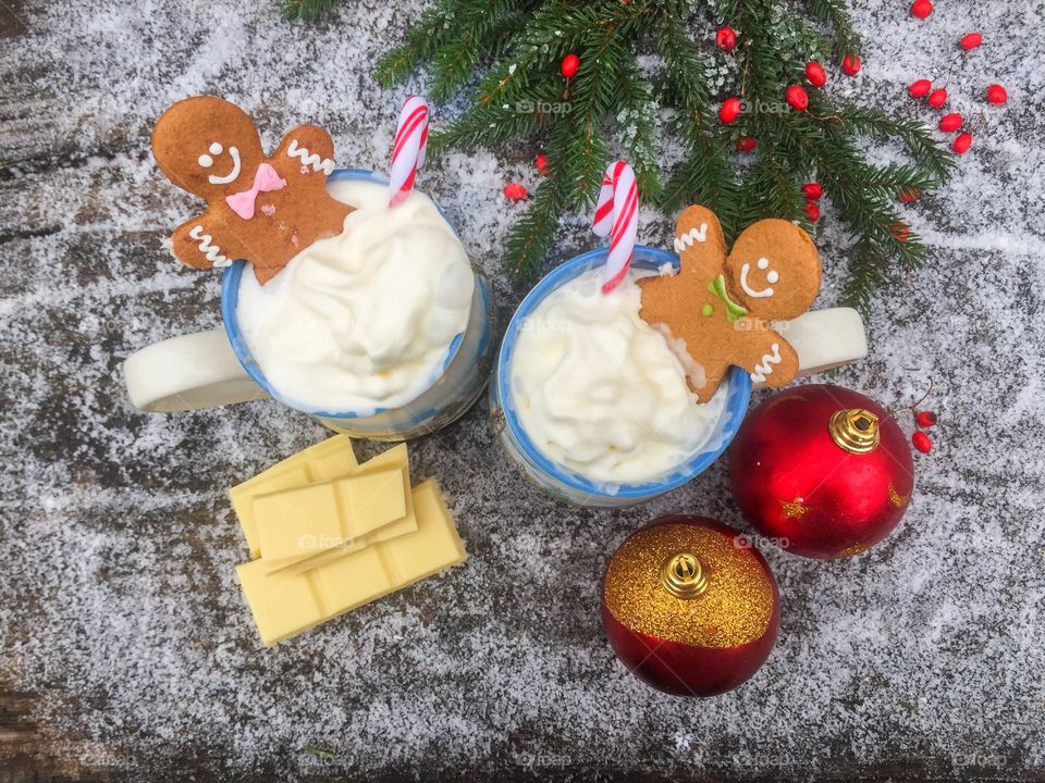 Two mugs of white chocolate with whipped cream, gingerbread man and candy canes on a snowy table with pieces of white chocolate and Christmas globes beside