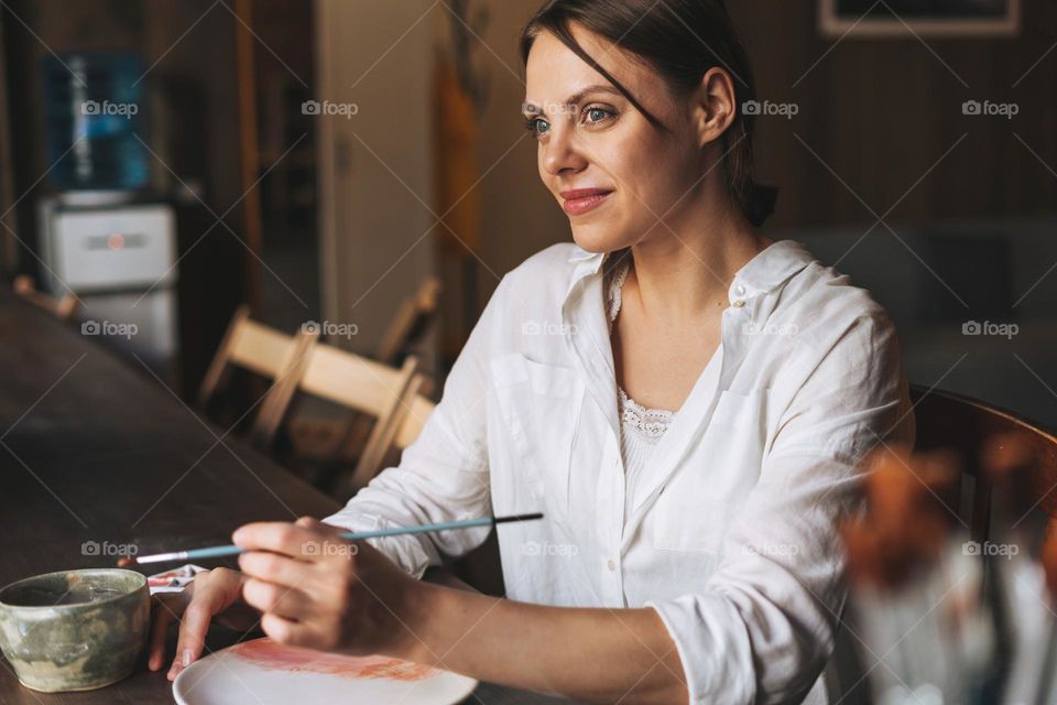 Young attractive woman in white shirt ceramic artist decorating clay plate with tool at table in pottery workshop. Handmade work student, freelance small business