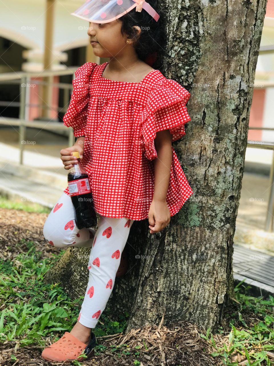 A little girl with red dress holding drink bottle and leaned over a tree.