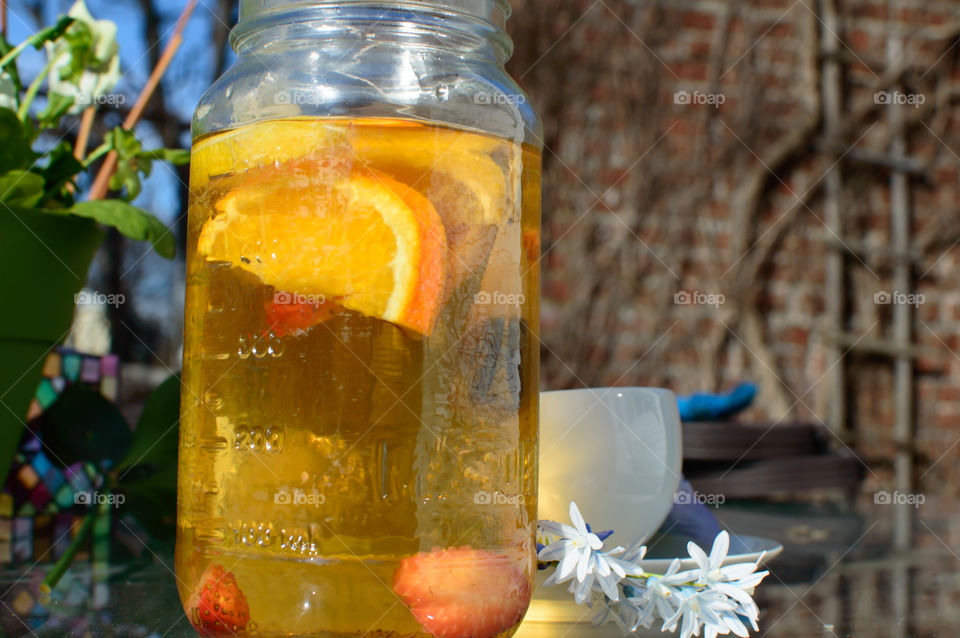 Making steeping fresh  herbal tea in glass jar with citrus and fruit using heat of the sun  outdoors on patio table with tea cup and flowers with blue sky and brick wall background 