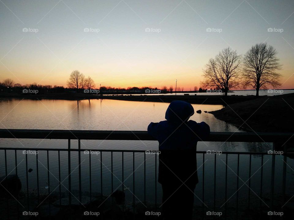 Boy leaning on railing with sunset over pond