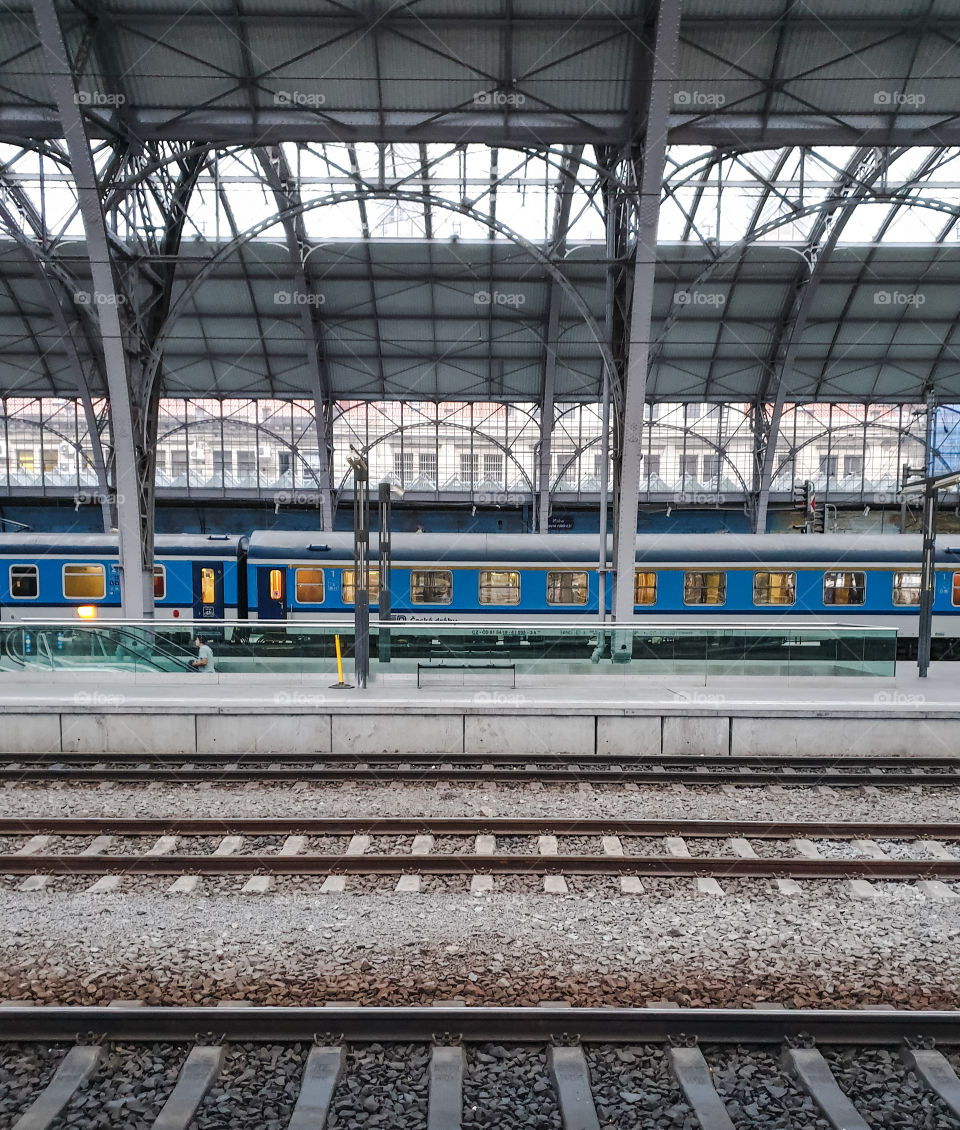 Prague, the main railway station.  A blue passenger train in the windows of which the light is on awaits boarding passengers on a platform with a glass roof