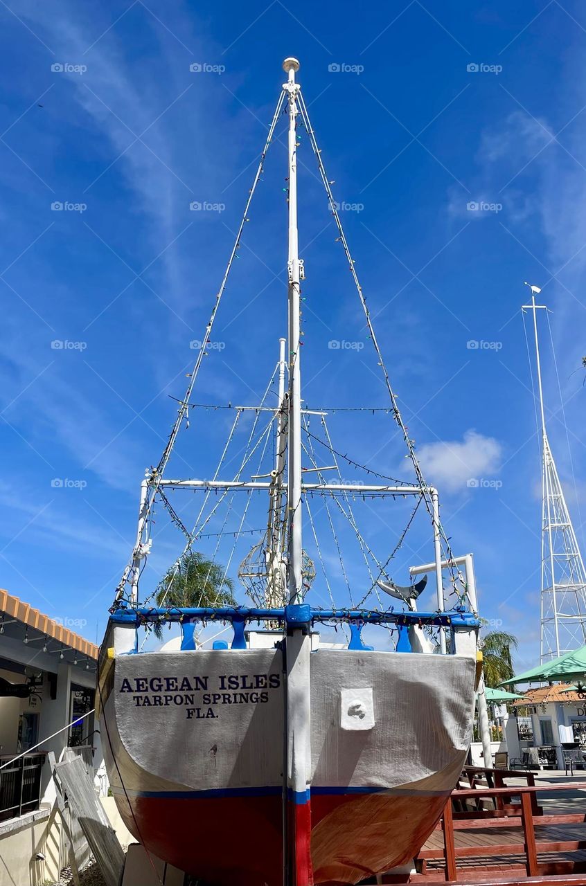 A vibrant fishing boat painted in red, white, and blue sits in the heart of Tarpon Springs, Florida. The boat stands out against the backdrop of a brilliant blue sky, with a few fluffy white clouds drifting lazily above.