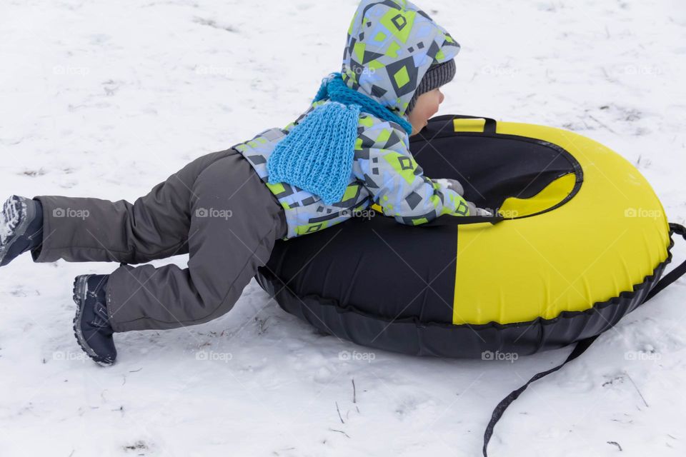A small, carefree boy walks in the white snow in winter and rides a tubing in the park, near trees in the snow.
