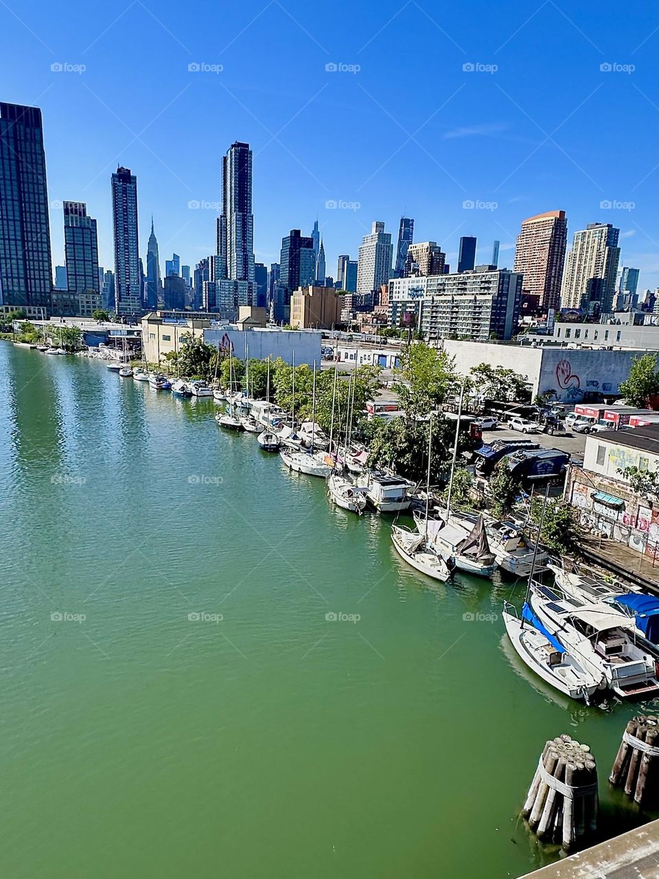 This is beautiful “Newtown Creek” with its many boats seen from the “Pulaski Bridge” that connects “Greenpoint”, Brooklyn to LIC, Queens. In the distance we see “Manhattan” including the “Empire State Building”. 2024. Hypnotic Productions