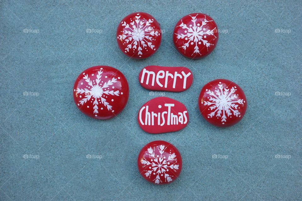 Merry Christmas text composed with red colored stones and hand painted stone snowflakes over green sand