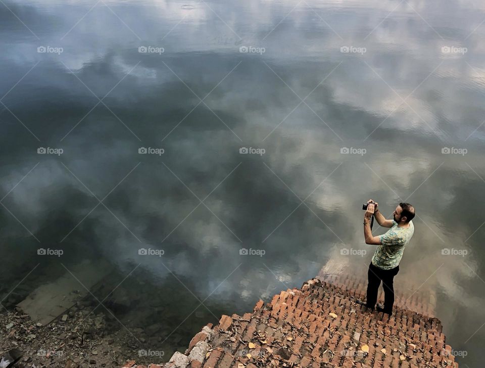 Steps descend downwards to the river where a man stands at the bottom taking a photograph, clouds are reflected in the water