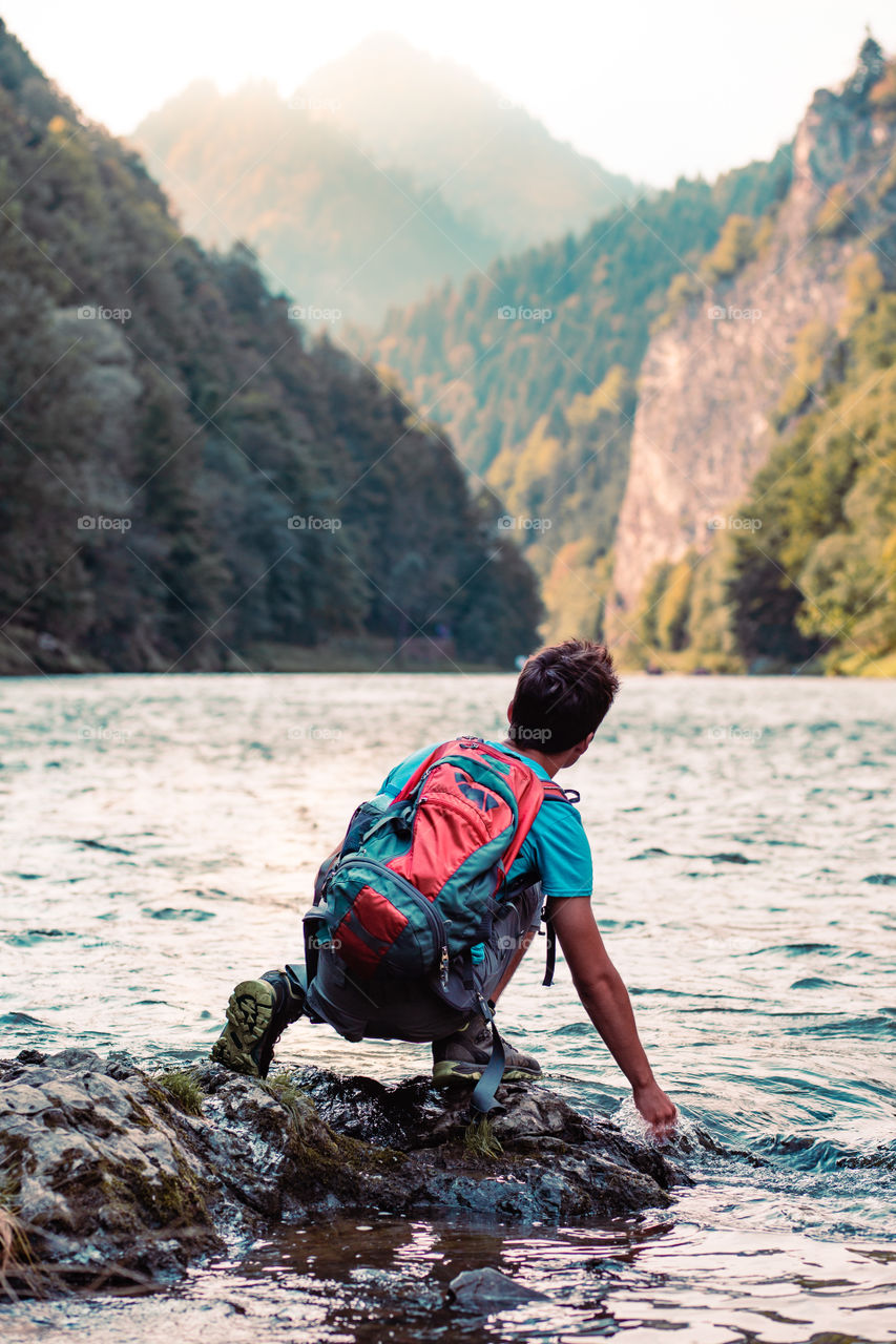Young boy with backpack sitting on rock over a river, holding hand in water, looking at mountains peaks, he is wearing sports summer clothes