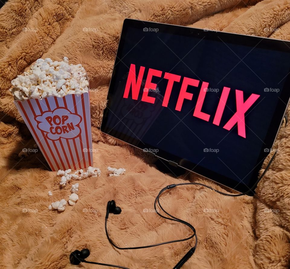 Laptop computer screen ready to watch Netflix for a fun, cozy, and relaxing night with popcorn as a snack, on soft bed blanket in room, with black earbuds