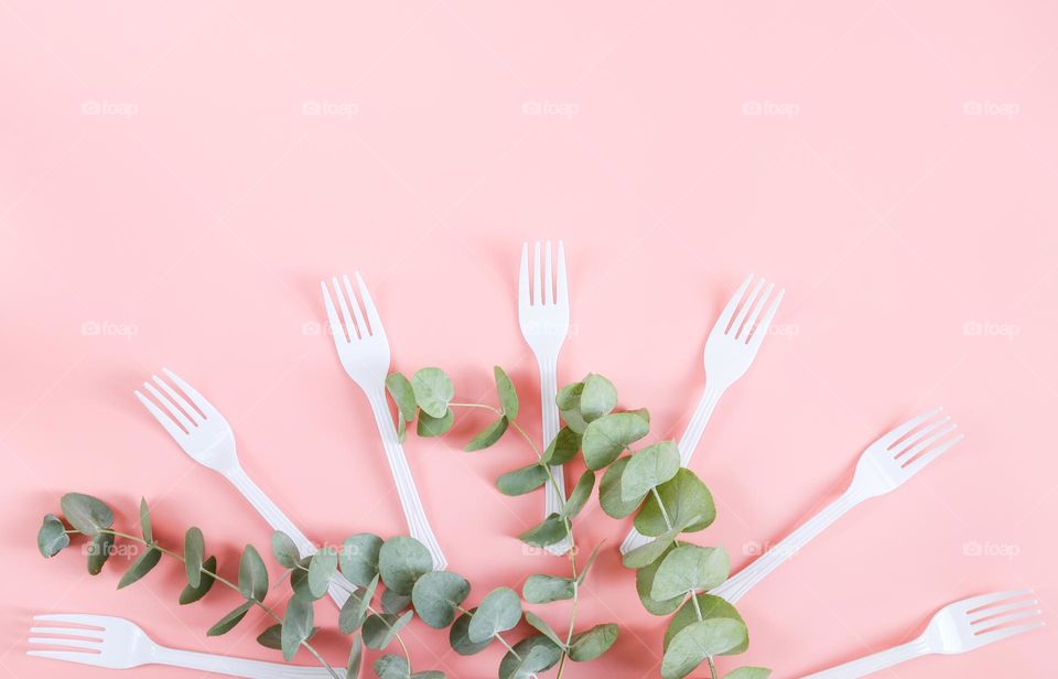 Seven plastic forks with eucalyptus branches lie on a pink ion with copy space, flat lay close-up. The concept of disposable tableware and ecology.