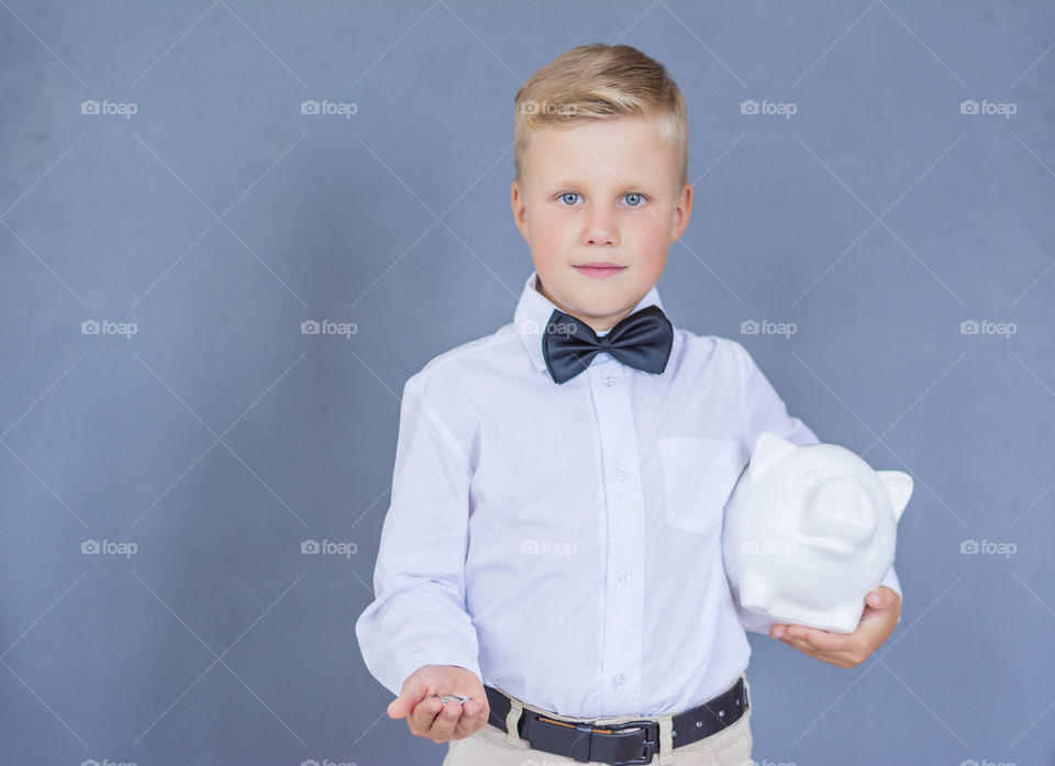 Beautiful smiling boy 6 years old in a white shirt with a butterfly stands in the studio on a blue background and shows holding coins in one hand, and in the other a piggy bank a white pig, thinking about investment bank