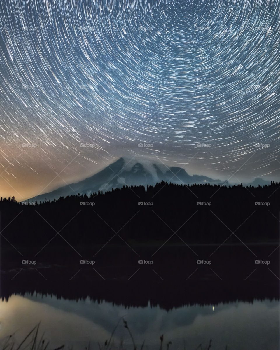 Star light trails over Mount Rainier on a dark night, with relfections in a still lake.