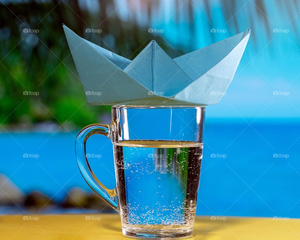a paper boat stands on a glass of water against the backdrop of the ocean