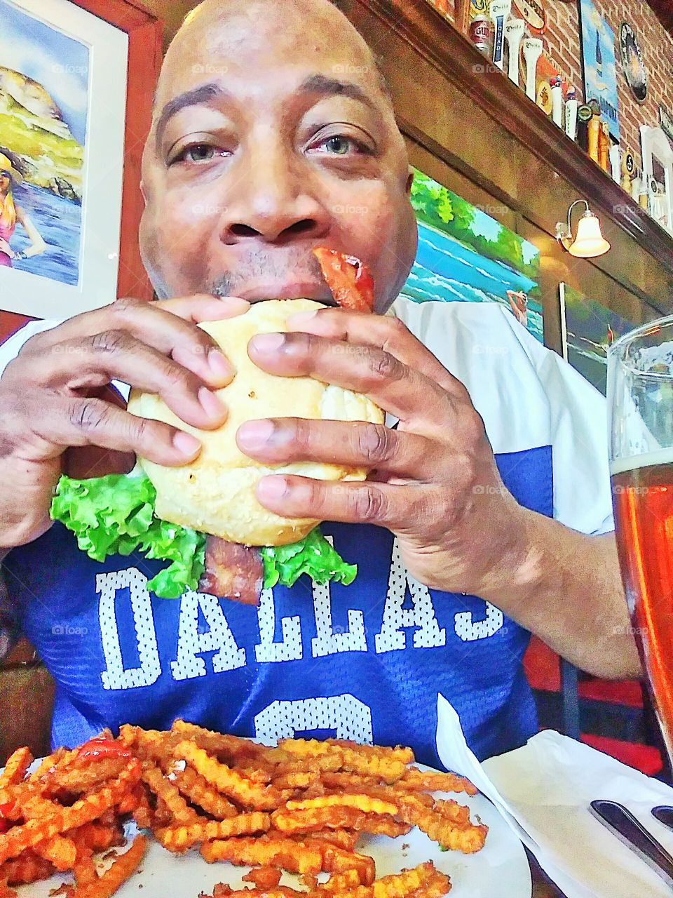 a man taking a big bite out of a avocado bacon burger with lettuce, onions, tomatoes, cheese and a side of sweet potato fries.