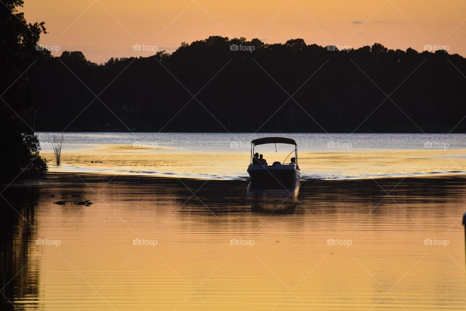 Boaters return to the lake shore at sunset. The orange sky and the trees are reflected in the water.