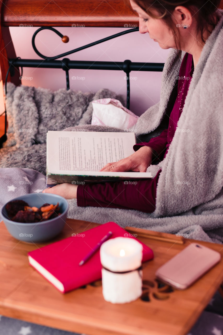 Woman enjoying the reading a book and drinking coffee at home. Young woman sitting in bed, wrapped in blanket, holding book, relaxing at home. Candid people, real moments, authentic situations