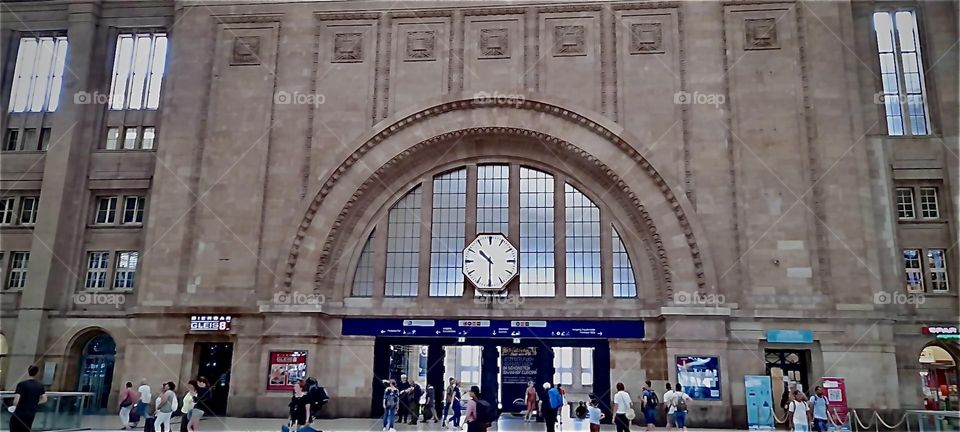 The train station of “Bad Ülster” in “Saxony” in Germany is seen from the outside with a large clock displayed inmidst the arched shape that is backed by semi transparent glass windows in between each structural partition. 2022. Hypnotic Productions