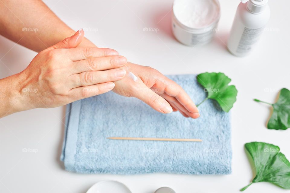 Woman applying  cosmetic moisturizing hand cream. Cosmetic products, green leaves on white table. Spa, manicure, skin care concept. Flat lay, overhead view