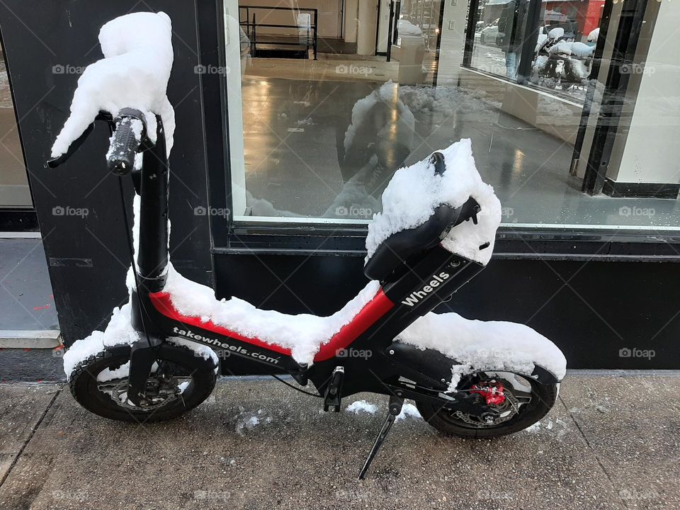 Little red motorcycle on the sidewalk half covered with snow.