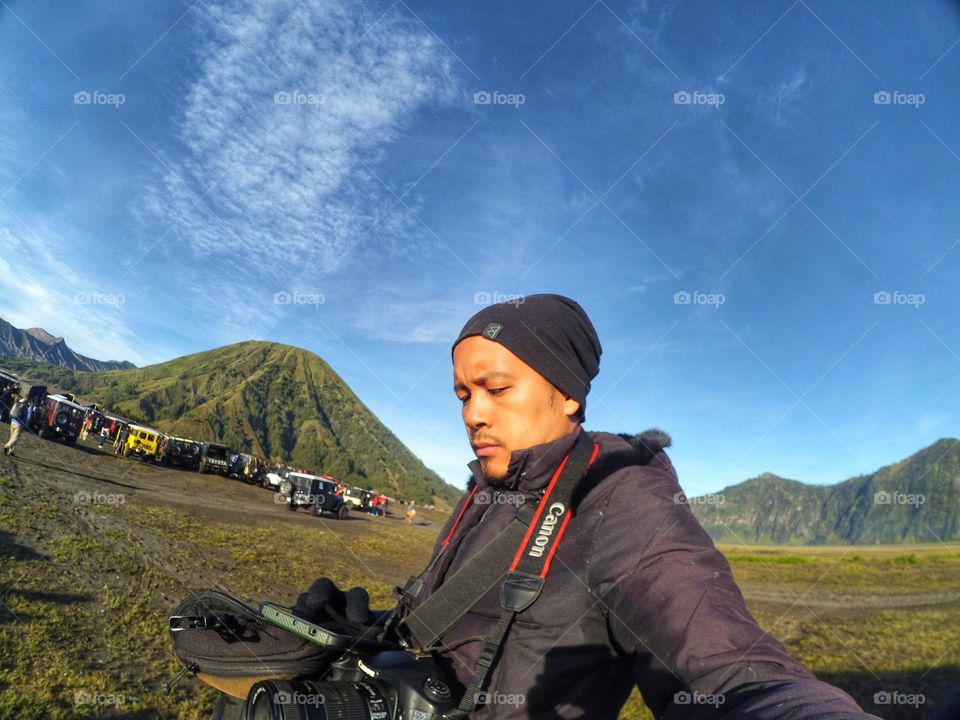 Selfie photo of a young Asian man in warm clothing against a green background in the Bromo mountains area with a cloudy sky. Vacation and adventure.