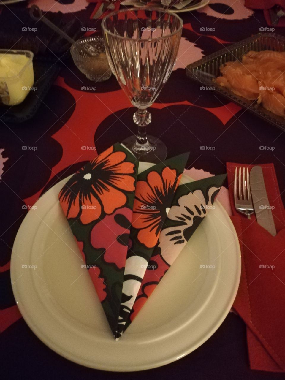 On a multicoloured table cloth a plate with a patterned napkin, a knife and a fork in a red pocket, a wine glass. In the background butter, mustard and gravlax on a green plate.
