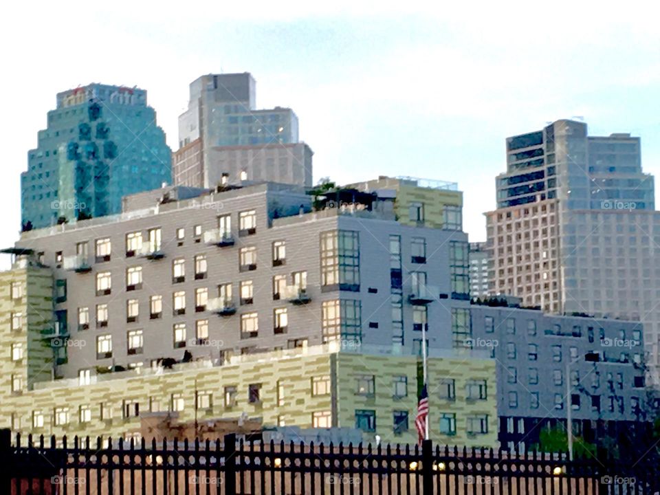 A Long Island City, Queens view of buildings of various sizes behind a railing near Borden Avenue by the waterfront of the East River. An American flag at half mast can also be seen in the foreground. 2020. Hypnotic Productions