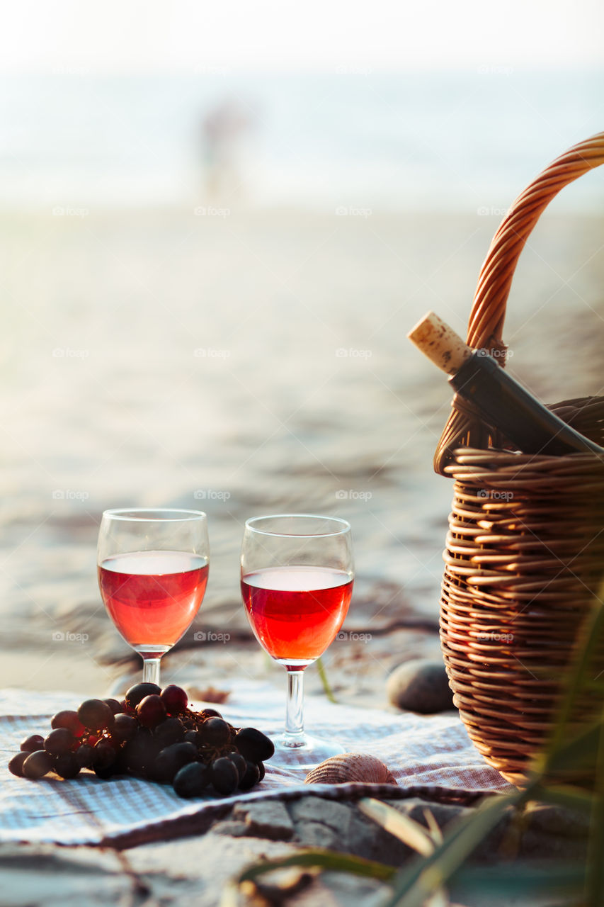 Two wine glasses with red wine standing beach, beside grapes and wicker basket with bottle of wine. Young couple standing in the sea in the background