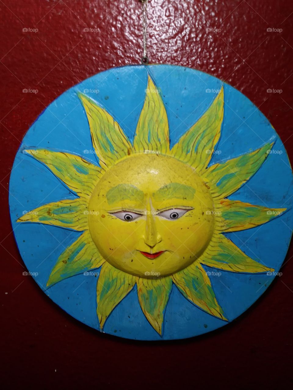 This is a painted plaster sun plaque hanging on a red wall. The sun is yellow and is surrounded by a blue circle. The sun is smiling.
