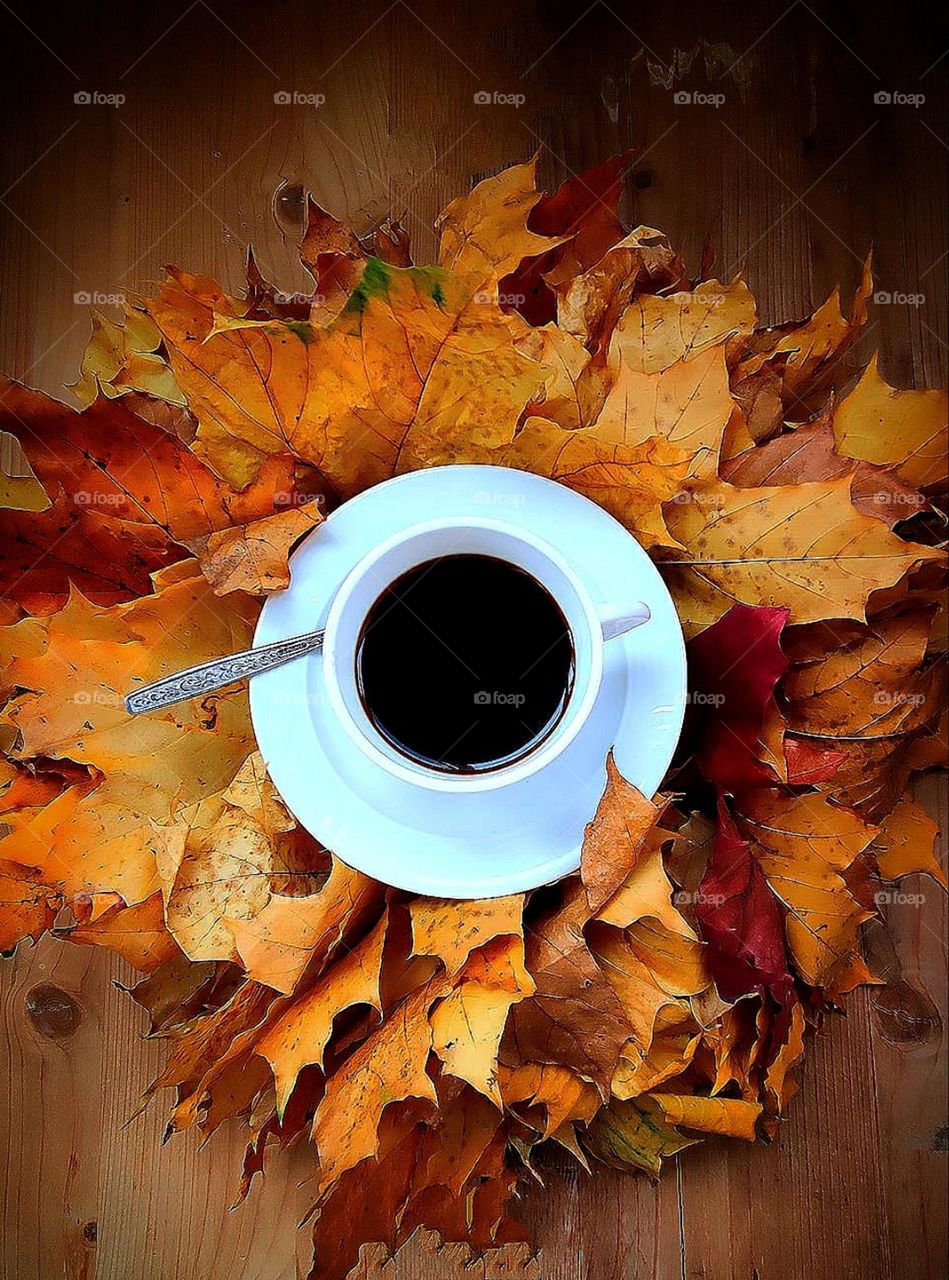 Autumn.  On yellow and red maple leaves is a white cup with black coffee.  Leaves look like tongues of fire