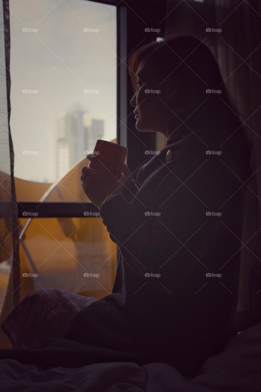 Asian woman sitting enjoying a cup of hot cocoa in front of a hotel room window in Singapore