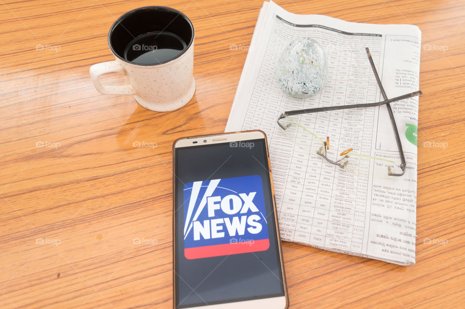 Kolkata, India, February 3, 2019: Fox news app (application) visible on mobile phone screen beautifully placed over a wooden table with a newspaper and a cup of coffee. A Technology Product Shoot.