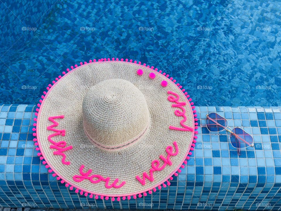 Summer hat with the words “wish you were here” and sunglasses on the edge of a pool with turquoise water