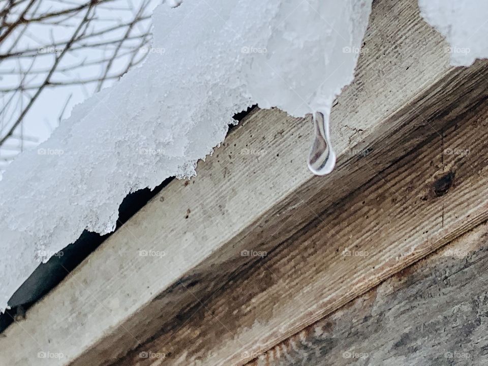 A drop of water falling from melting snow and ice on the roof overhang of a wooden structure 