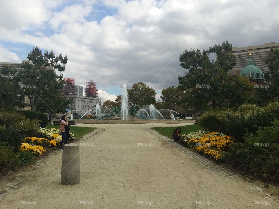 Off center picture of a park on a cloudy day. It features a fountain and lots of gree greenery