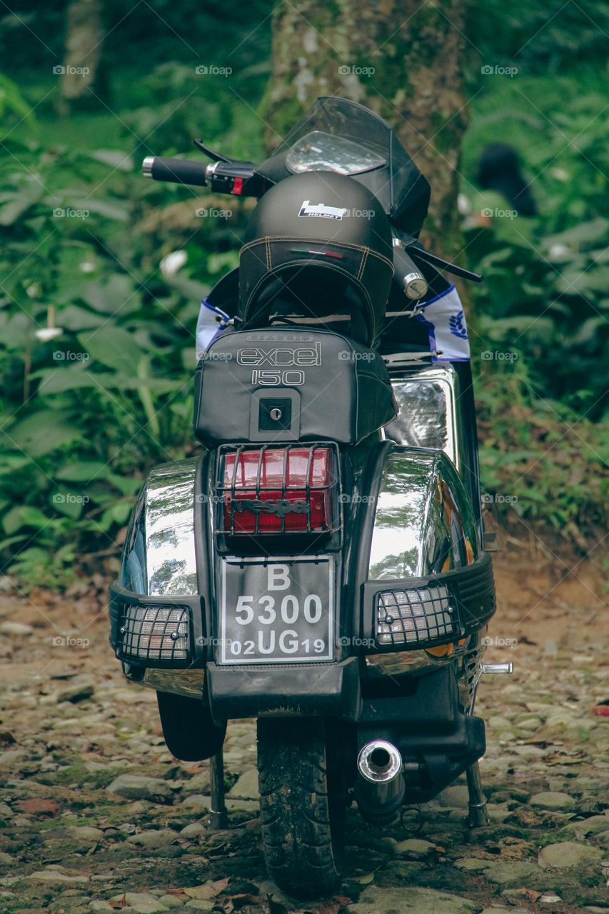 Back view of a classic vespa type excel 150 with a 2 stroke engine with a capacity of 150 cc, with chrome side bodies. against a backdrop of green trees.