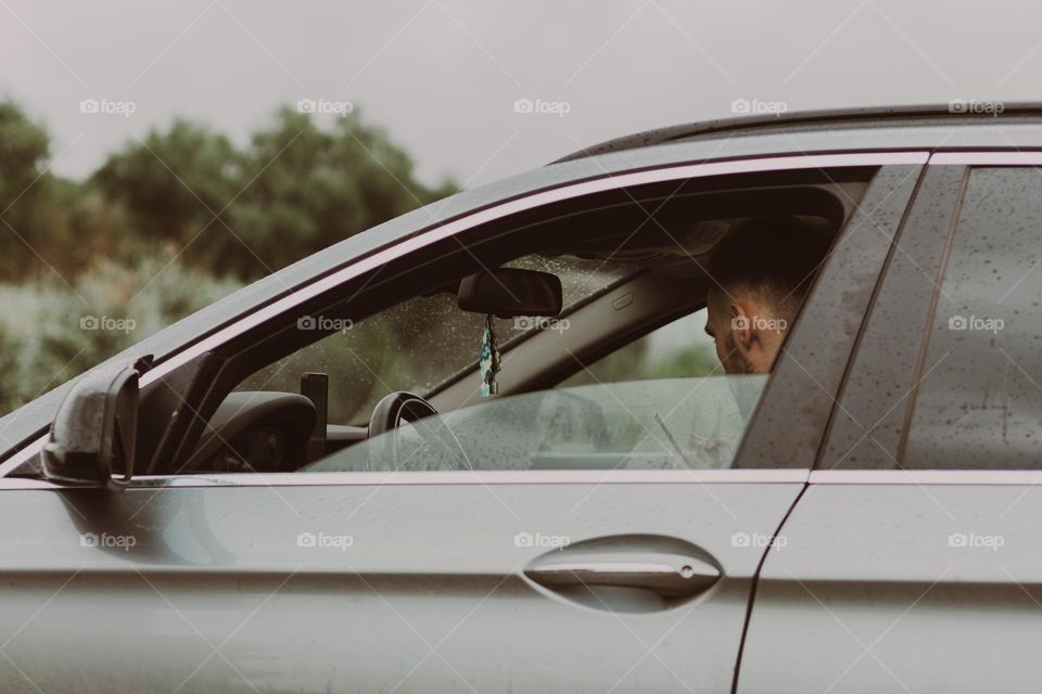 Portrait of one Caucasian unrecognizable guy sitting behind the wheel of his car, turning his head to the side, on a rainy cloudy day with the glass down on the door, close-up side view.