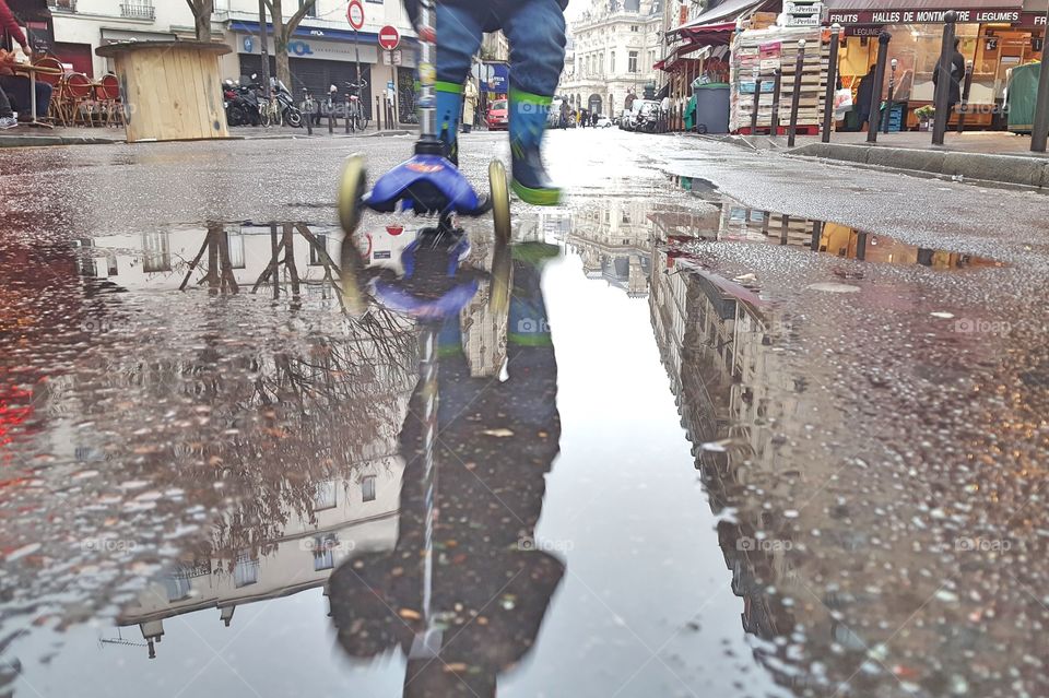 child on a scooter reflection in a puddle of water. The toddler is wearing rubber boots in the street in Paris