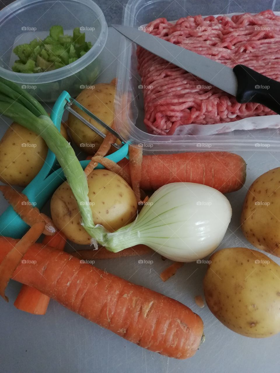 Fresh ingredients of a minced meat soup on a grey table. The meat in a plastic box on a paper,knife is on them. Uncut, not peeled potatoes and onion, partly cut, peeled bumpy carrots, celery in a cup on a scratched chopping board.