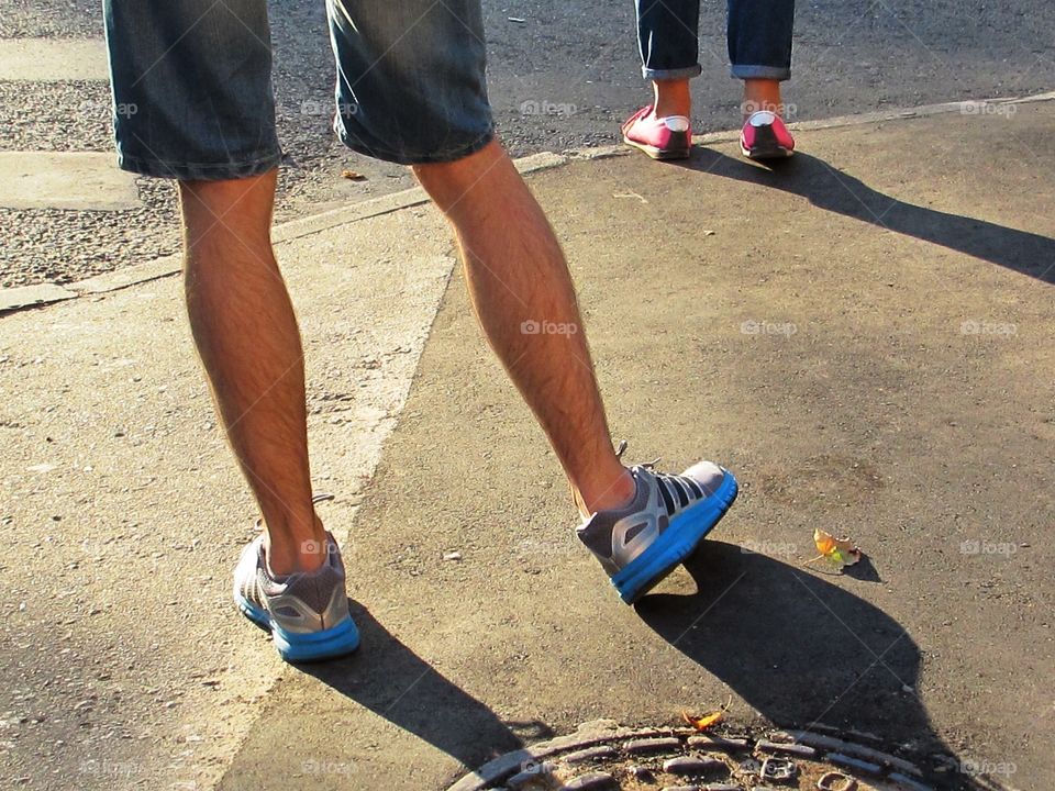 Man and woman feet outdoors. The guy is standing behind the girl. The pic only legs in jeans and pink and blue sneakers 