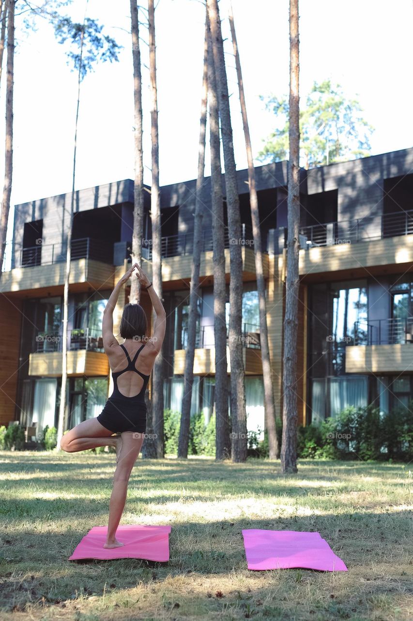 a girl in a black tracksuit, on a pink rug in the forest, does yoga, the girl meditates