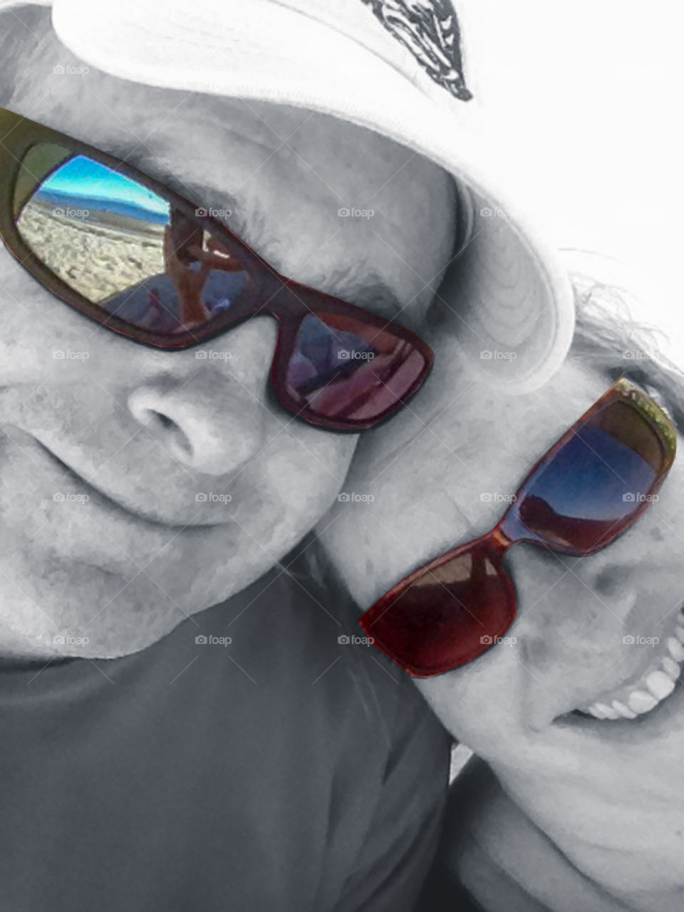 A selfie of a husband & wife at the beach. The photo is primarily B&W except for the brown sunglasses and reflections. The reflections show; the sandy beach,  the iphone, body parts & the ocean, mountains & sky in varying shades of beautiful blue! 💙