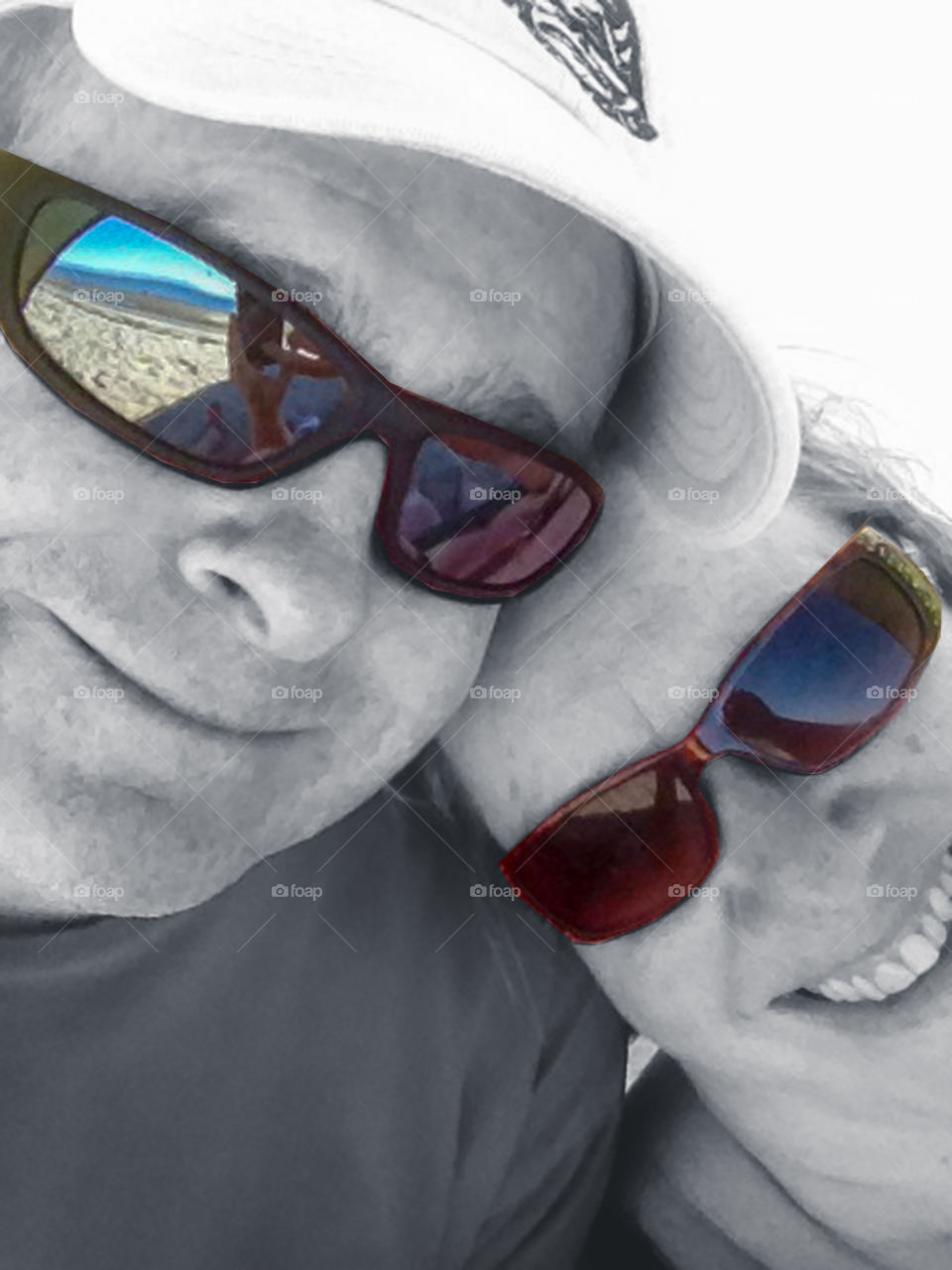 A selfie of a husband & wife at the beach. The photo is primarily B&W except for the brown sunglasses and reflections. The reflections show; the sandy beach, the iphone, body parts & the ocean, mountains & sky in varying shades of beautiful blue! ๐