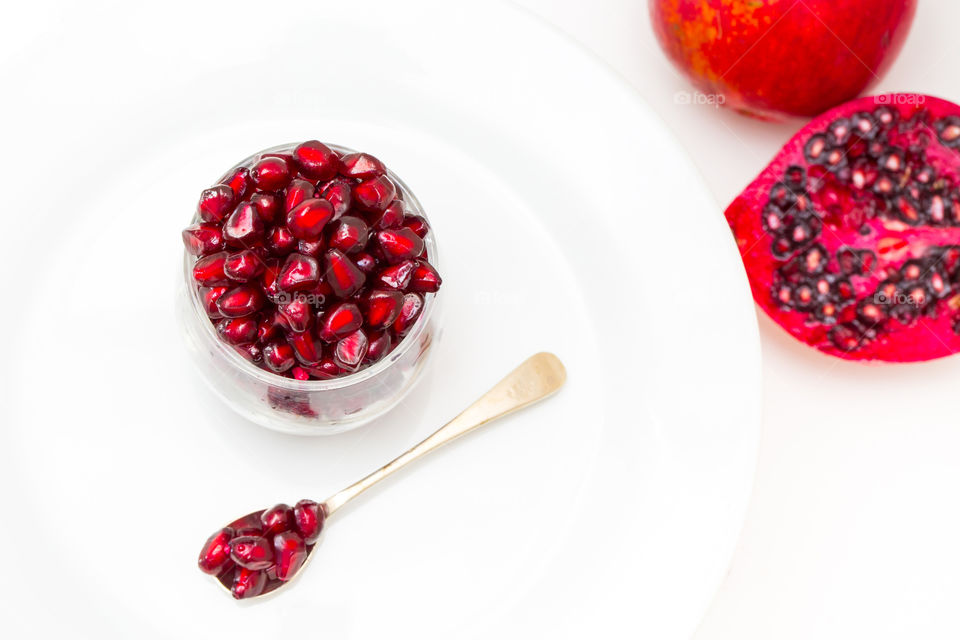 Pomegranate seeds with sliced fresh  pomegranate. Served on a white plate with a single spoon. Close up flat lay with a white background. Fresh summer fruit.