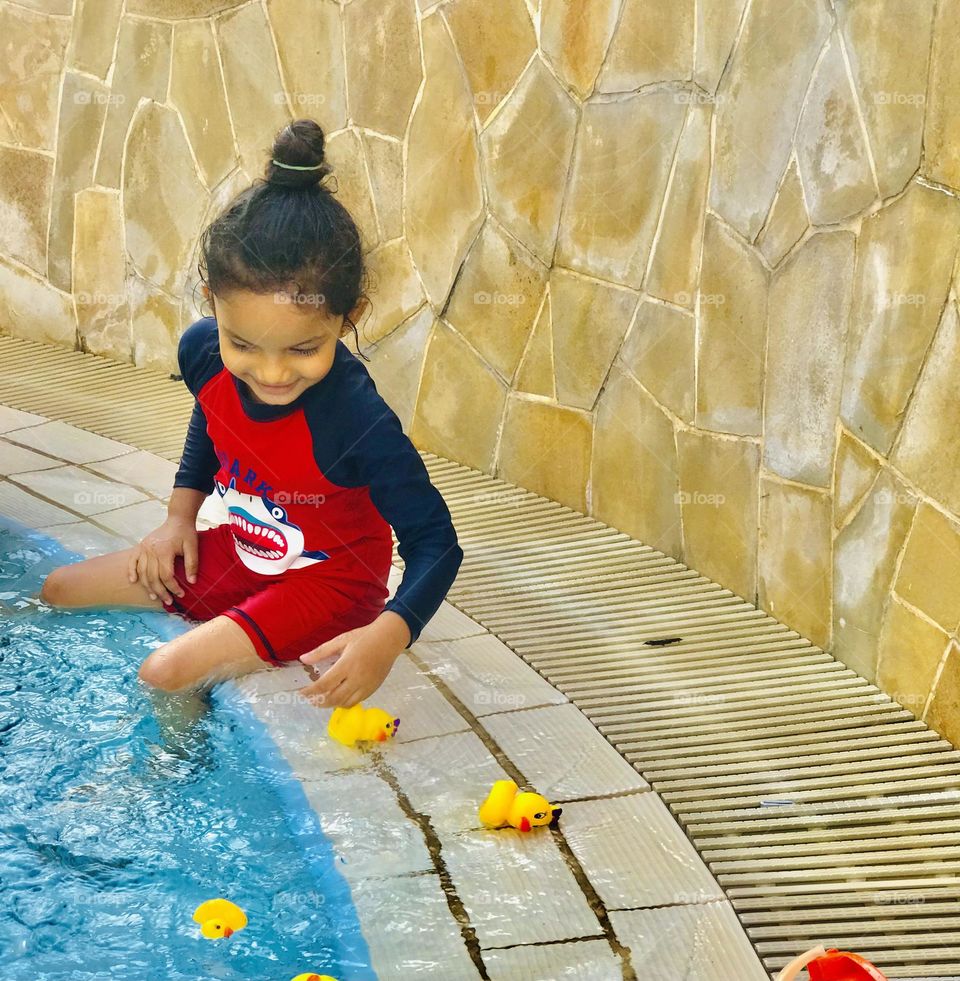 A 4 year old baby girl wearing red and black swimsuit and she put her legs into the swimming pool and playing with yellow ducks and gives great smile 😊