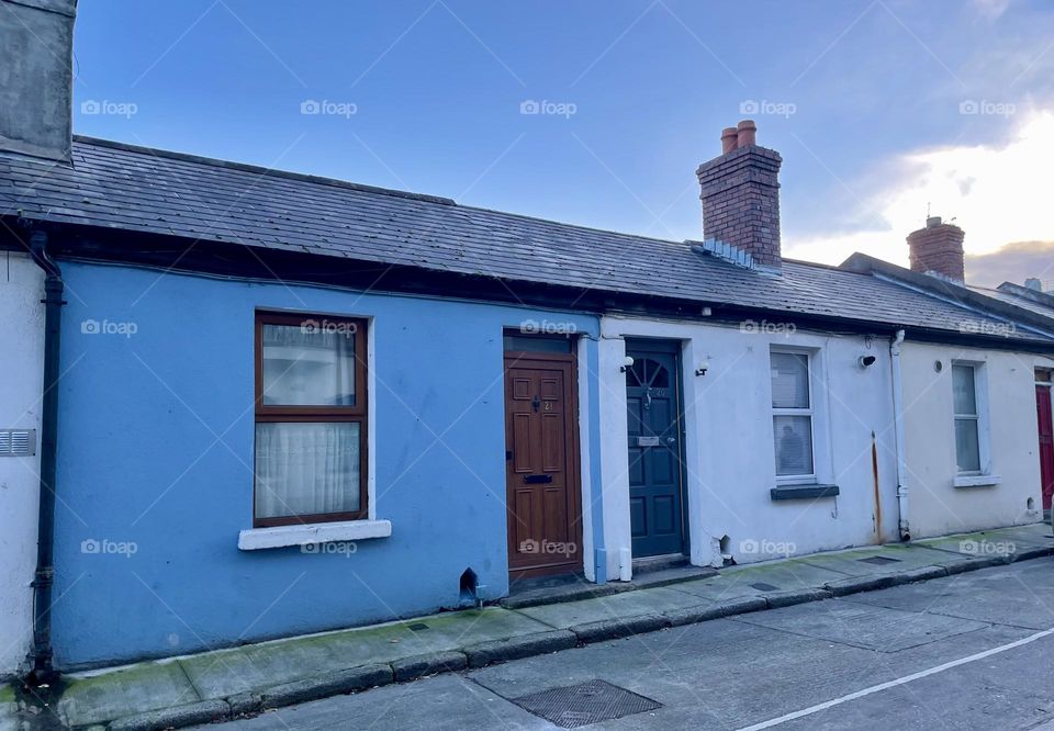 A row of small, colorful attached one-story Irish houses lining a peaceful street in Dublin. Each house, with its low ceilings and quaint, compact design are on a quiet street, free of hustle and bustle, adding to the almost nostalgic atmosphere.